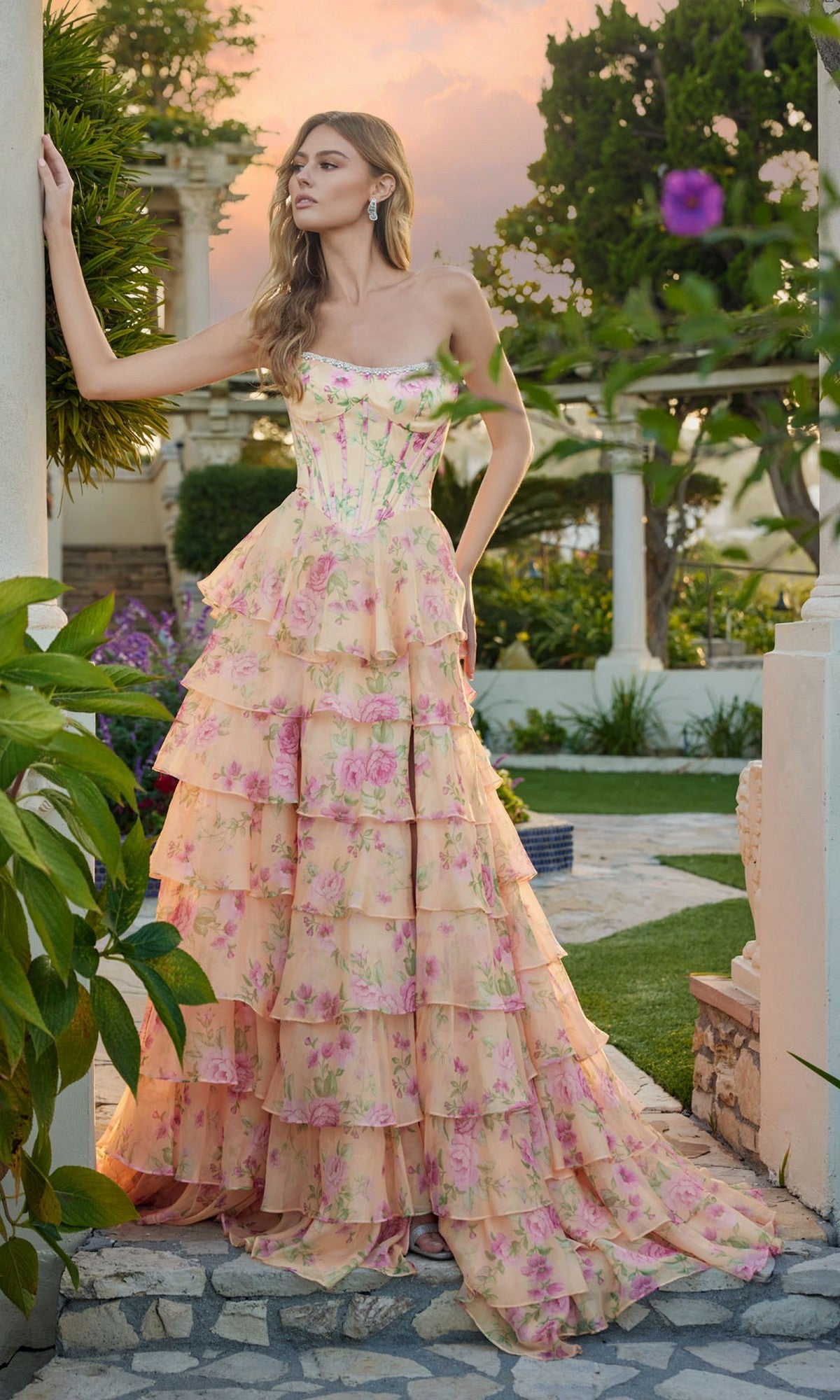 At sunset, a woman stands outdoors in the Juliet JT2739S long prom dress, featuring a strapless floral corset bodice and flowing tiered chiffon A-line skirt. She rests her hand on a column and gazes to the side.